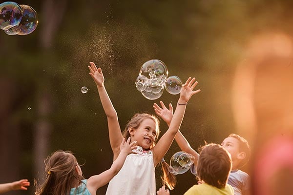 One smiling girl in a group of kids jumps up into the air catching glistening bubbles.