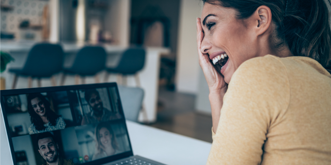 woman sitting in her kitchen laughing in front of a computer with coworkers on video call