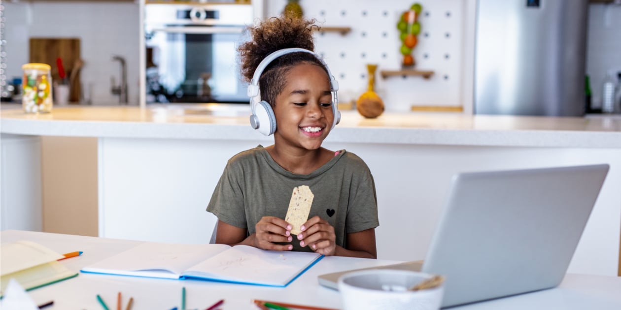 photo of young girl sitting in the kitchen wearing headphones and looking at a laptop with papers and colored pencils