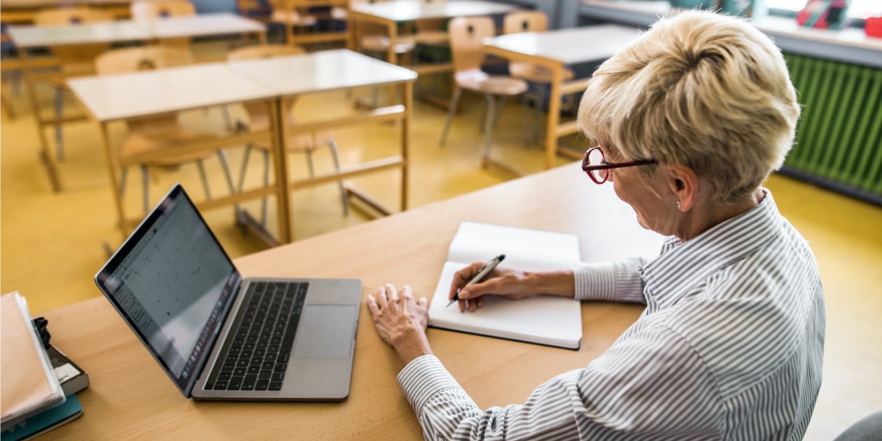 photo of older teacher sitting at her desk in empty classroom and taking notes next to a laptop