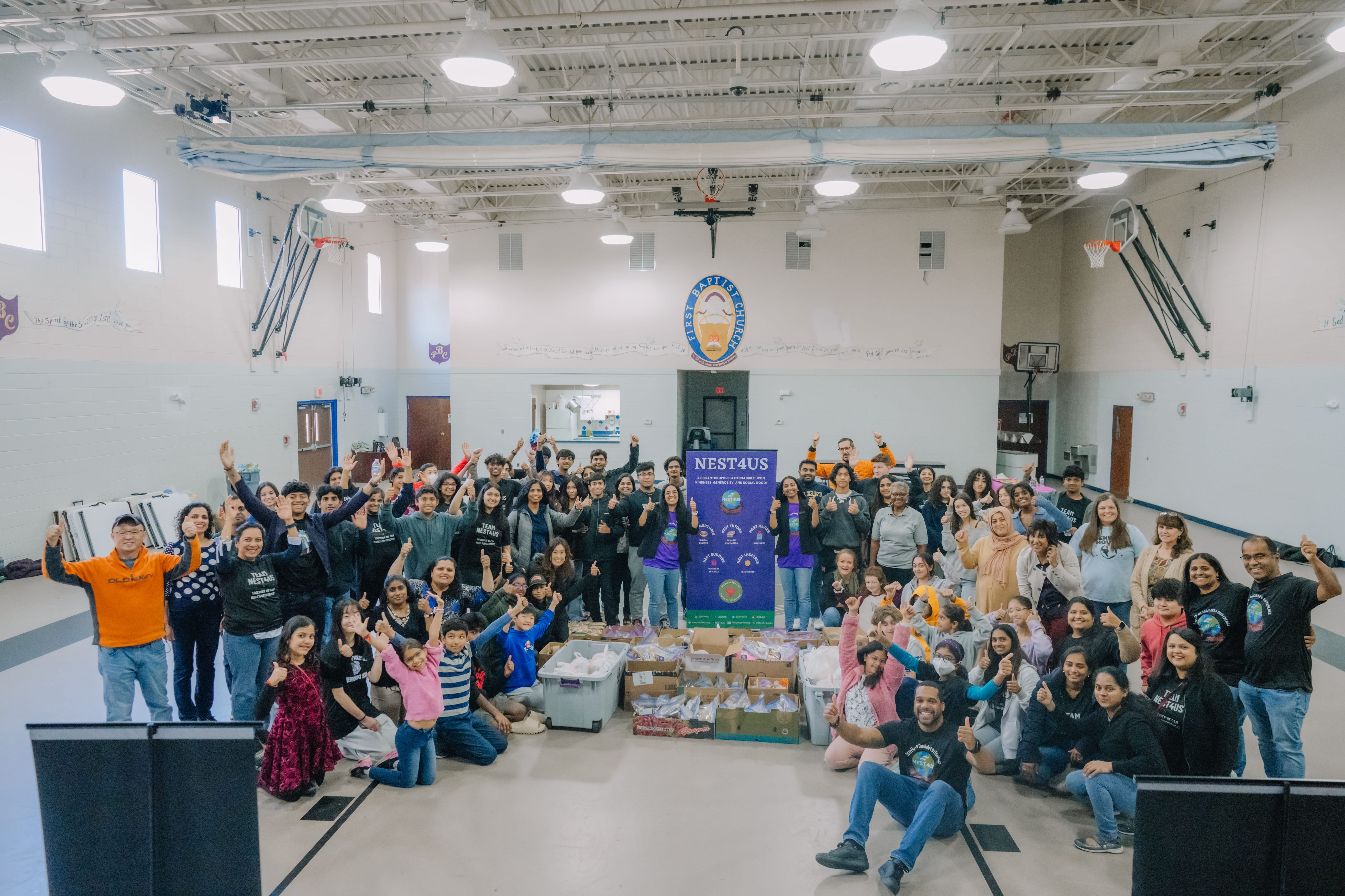 Dozens of NEST4US volunteers of all ages cheering around a NEST4US banner and boxes of packed Thanksgiving bags in a gym